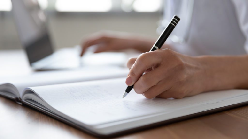 Female doctor hand making notes in journal using laptop